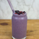 A peanut butter & jelly smoothie in a glass cup with a glass straw, with mixed frozen berries on top.