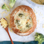 Broccoli Cheddar Soup in a bread bowl with part of the pot showing and a wooden spoon with shredded cheese on it.