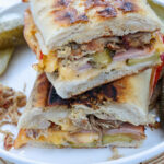 Two cuban sandwiches stacked on top of each other on a white plate with pickles on the side.