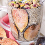 Clear glass mug with chia seed pudding, lined with fig slices, topped with pistachios and black sesame seeds.
