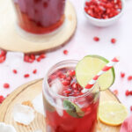 Pomegranate fizz mocktail in a clear glass with red and white striped paper straw, lime garnish and a pitcher of the drink in the background.